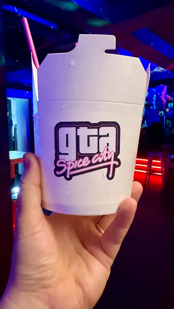 A promotional video for NQ64, a retro arcade bar in Cardiff. The video showcases the venue's neon-lit atmosphere, with clips of people playing various arcade games, including racing and shooting games. It also features close-ups of bartenders preparing several colorful cocktails, such as the 'Jurassic Marg', 'Aku Aku fruit punch', and a sharing drink called 'GTA Spice City' served in a white takeaway box with fortune cookies.