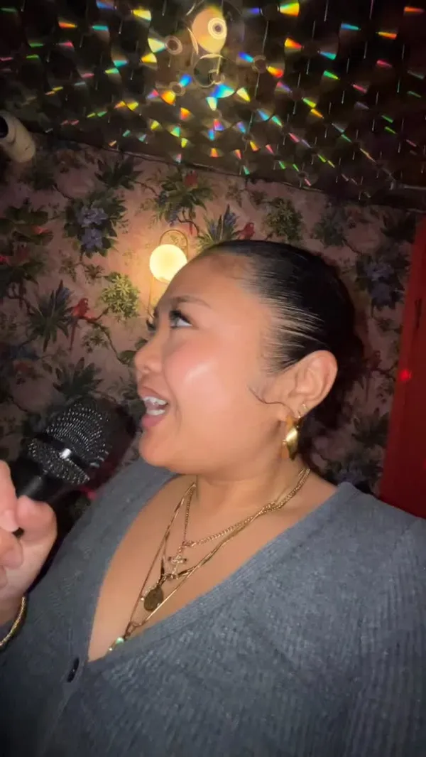 A group of friends are at a karaoke bar. The video shows them singing in a private karaoke pod, which has floral wallpaper and a ceiling decorated with CDs. It also includes quick shots of the main bar area, which has neon signs, disco balls, and a well-stocked bar. The group is shown eating pizza and drinking various cocktails at a booth. The overall vibe is energetic and fun, focusing on the social experience of karaoke and drinks with friends.