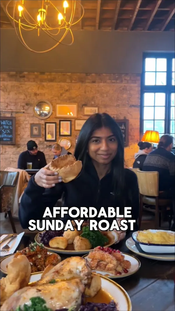This video showcases a Sunday roast at Forest Tavern, a gastropub located near Forest Gate. The video features a woman sitting at a table laden with multiple plates of food, including a half chicken, roast beef, Yorkshire puddings, roast potatoes, vegetables, mac and cheese, and fried halloumi with pomegranate seeds. The food is presented in a rustic, hearty style. The main focus is on highlighting the generous portions and affordable price of the Sunday roast.