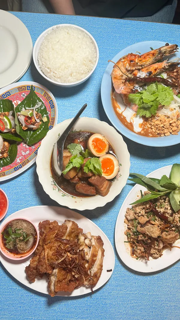 The video promotes a Thai food pop-up located inside a pub in London. It showcases a large spread of authentic Thai dishes, including Hat Yai fried chicken with fried shallots, Larb Moo (spicy minced pork salad), Kai Palo (pork belly and egg stew), Miang Kham (leaf-wrapped bites), and grilled prawns. The creator is featured enjoying the meal, and the voiceover emphasizes the chef's Thai heritage and how the food evokes memories of Bangkok.