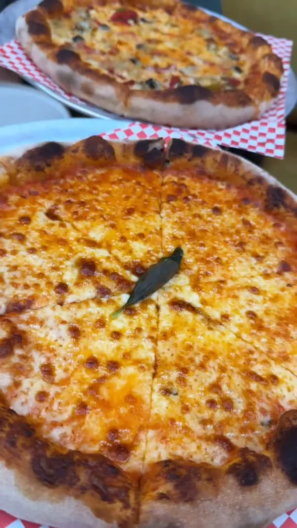 The video promotes a New York-style pizza pop-up, 'Proper Slice at Eve', located within the Ember Locke hotel in Kensington, London. It showcases the venue's stylish interior and pleasant outdoor garden seating. The food featured includes a burrata with pesto appetizer, a classic cheese pizza, and a vegetable-topped pizza. Key moments include a close-up of the burrata being cut, a satisfying cheese pull from a slice of veggie pizza, and dipping the pizza crust into a ranch sauce. The creator is shown enjoying the meal throughout the video.