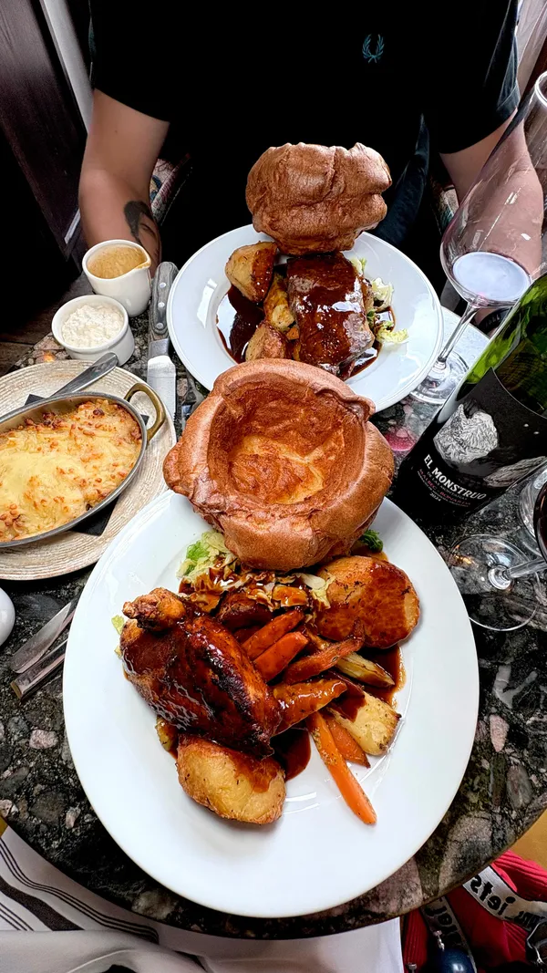 The video showcases roast dinners, including chicken and beef options, served with large Yorkshire puddings, roasted vegetables, and potatoes. It features close-ups of gravy being poured over the meat and chicken, a shot of cheesy cauliflower, and a sticky toffee pudding dessert with ice cream and sauce. The video also includes interior shots of the pub/restaurant, showing the seating area and bar, and an exterior shot of the entrance with 'THE GUN' sign.