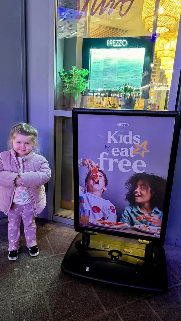 This video promotes Prezzo, an Italian restaurant, highlighting their "Kids Eat Free" promotion. The video starts with an outdoor shot of the restaurant's signage and outdoor seating area. It then shows a poster advertising the promotion, followed by a family entering the restaurant. The video showcases the kids' menu, a young girl coloring at the table, and various food items, including garlic bread, pizza with pesto and tomatoes, fried calamari, a burger with fries, and rigatoni pasta with tomato sauce. The presentation of the food is casual and family-friendly. The video concludes with the young girl enjoying a sundae. There is no specific dietary information mentioned, but the menu shown includes vegetarian options. The main focus is on showcasing the restaurant's kid-friendly atmosphere and the "Kids Eat Free" promotion.