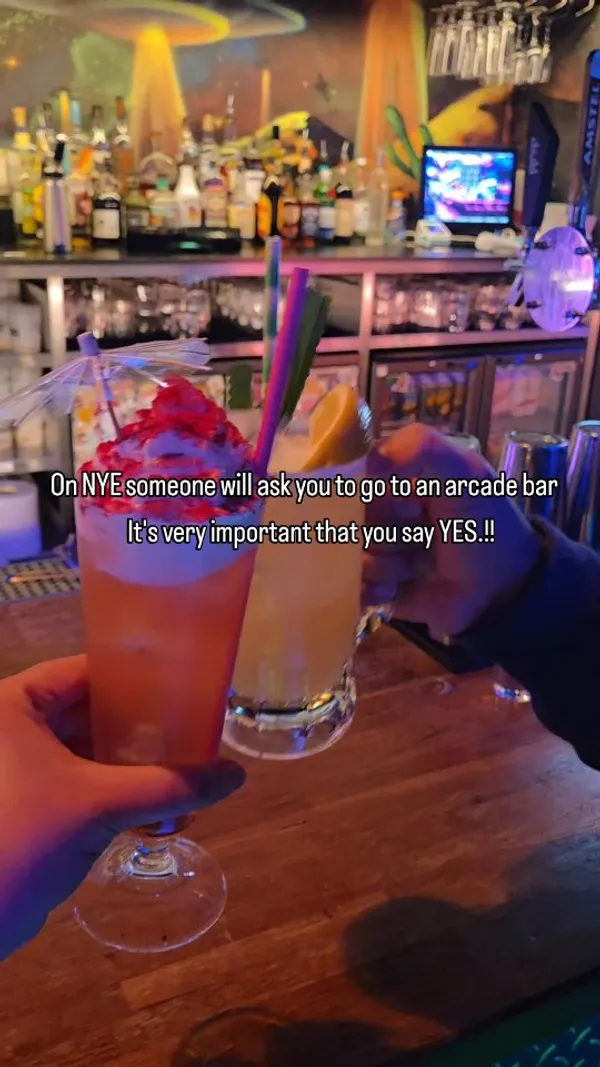 The video showcases an arcade bar called NQ64. It begins with a wide shot of the dimly lit, neon-heavy interior, filled with people. It then cuts to a close-up of two colourful cocktails being cheered. The rest of the video features various classic arcade games like Mario Kart, Tekken, and Time Crisis, with people actively playing them. The decor is highlighted, featuring UV-painted walls and a unique ceiling covered in posters. The overall atmosphere is portrayed as vibrant, fun, and energetic.