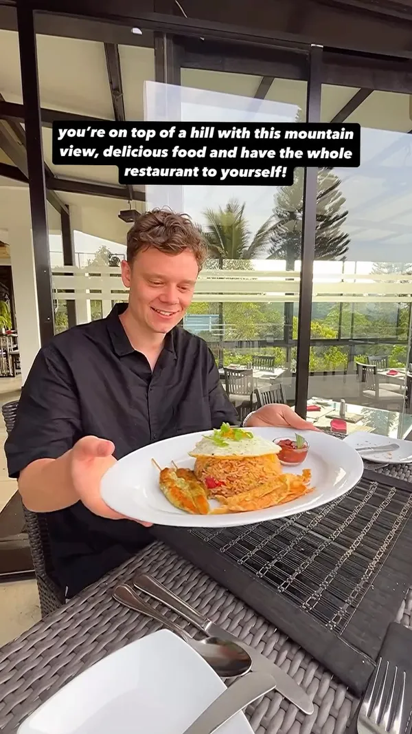 The video showcases Theva Cuisine restaurant in Kandy, Sri Lanka, highlighting its hilltop location with views of the Hanthana mountains. It features a man presenting a plate of nasi goreng topped with a fried egg, chicken skewers, crispy wonton strips, and a side of chili sauce. A woman is later seen enjoying Sri Lankan fish curry with rice and other accompaniments. The video also shows the restaurant's outdoor seating area, a swimming pool, and the surrounding lush greenery. The main focus is on the restaurant's scenic location and the presentation of its Sri Lankan cuisine.
