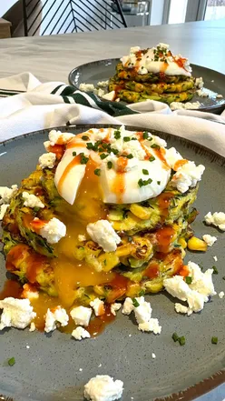 Savory courgette and sweetcorn pancake stack topped with feta, poached egg, and chili sauce.
