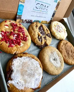 Box of cookies from CRM Cookie Co featuring raspberry ripple, chocolate chip, birthday cake, cinnamon roll, sugar, and spiced biscoff cookies.