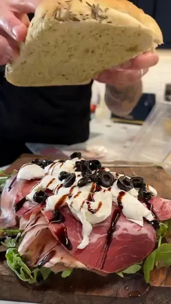 Italian focaccia sandwich with pesto mayo, rocket, meats, burrata, balsamic glaze, and olives, perfect for summer dinner.
