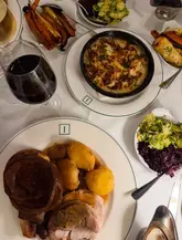 An overhead flat-lay shot of a Sunday roast dinner spread at a restaurant, featuring a plate with roast turkey, potatoes, and a Yorkshire pudding, alongside side dishes like cauliflower cheese and roasted vegetables, with a glass of red wine.