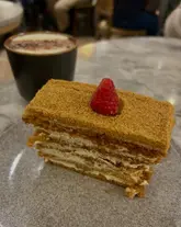 A close-up, eye-level shot of a slice of honey cake served on a grey plate, with a Hojicha latte visible in the background. The image captures a casual cafe setting, focusing on the layered texture of the cake.