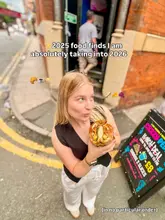 A high-angle, outdoor selfie of a creator holding a large sandwich, serving as a cover image for a 'food finds' listicle video. The image features a street-level background with a sandwich board sign, capturing a casual, on-the-go dining vibe.