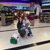 A candid family photo taken in a cinema lobby, featuring a mother and her two children posing in front of a candy machine and merchandise displays. The image captures a casual family outing moment.