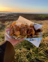 A handheld, close-up shot of deep-fried pigs in blankets topped with gravy, served in branded paper, captured against a scenic Scottish beach at sunset.