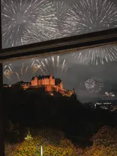 A night-time photograph taken from an interior window looking out at Edinburgh Castle, which is illuminated against a dark sky filled with fireworks. The image captures a celebratory, high-end atmosphere typical of a premium hospitality venue during New Year's Eve.