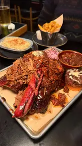 A fast-paced montage showcasing a large BBQ meat platter, sides like mac and cheese and onion rings, and cocktails at a restaurant. The video uses quick cuts and upbeat music to highlight the food.