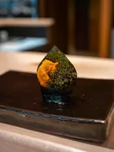 A high-quality, close-up shot of a piece of sushi, specifically a gunkan maki topped with uni and caviar, served on a dark, minimalist plate. The lighting and focus are expertly handled, highlighting the textures of the ingredients, which is characteristic of high-end omakase photography.