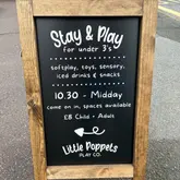A clear, well-lit photograph of a wooden A-board sign advertising a 'Stay & Play' session for children under 3 at Little Poppets Play Co. The sign provides essential details including activities, times, and pricing.