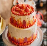 A vibrant, two-tier wedding cake featuring an abstract, textured frosting design in shades of orange, pink, and white, accented with gold leaf and piped rosettes. The cake is presented on a stand, though the composition suffers from tight framing that clips the top of the cake topper.