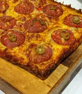 A close-up, high-angle shot of a homemade Detroit-style pizza served on a wooden board. The pizza features a thick, crispy crust with pepperoni and jalapeño toppings. The lighting is bright and direct, emphasizing the texture of the cheese and the oily sheen of the pepperoni, though the composition feels somewhat tight and cropped.