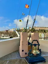 A high-quality, well-composed photograph taken from the deck of the SS Great Britain in Bristol. The image features the ship's bell in the foreground with the iconic colourful houses of the Clifton/Hotwells area in the background, capturing a bright, sunny day.
