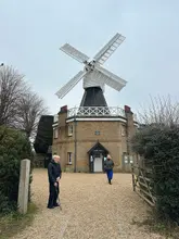 A candid, wide-angle snapshot of an elderly man and a woman standing in front of the Wimbledon Windmill museum on a grey, overcast day. The image functions as a personal travel memory rather than a polished marketing asset for the venue.