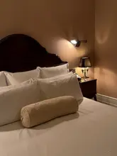 A moody, atmospheric shot of a hotel bed at The Ned London, focusing on the warm, inviting lighting and clean, well-made bedding. The image captures a sense of luxury and comfort, typical of a high-end hotel stay.