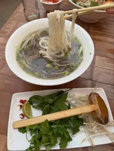 A top-down/45-degree shot of a bowl of beef Pho being lifted with chopsticks, accompanied by a side plate of fresh herbs, bean sprouts, lime, and chili on a wooden table.