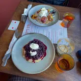 A top-down, overhead shot of two plates of pancakes served on a wooden table. The scene includes cutlery, napkins, a menu, and a playing card, capturing a casual brunch setting.