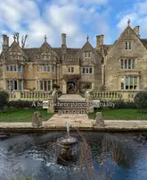 A wide, eye-level shot of a historic stone manor house hotel, featuring a fountain in the foreground and a clear blue sky with clouds. The image is used to promote a family-friendly hotel experience.