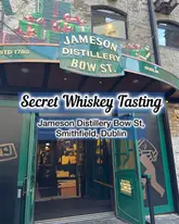 A straight-on, eye-level shot of the entrance to the Jameson Distillery on Bow Street in Dublin. The image features the prominent green signage above the doorway, with text overlays indicating the location and the 'Secret Whiskey Tasting' experience.