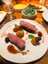 A high-angle, close-up shot of a restaurant table featuring a main course of duck breast with a croquette, accompanied by side dishes including mashed potato, creamy spinach, and tender stem broccoli. The image captures the ambient, warm atmosphere of the venue, though the lighting is heavily skewed towards orange tones.