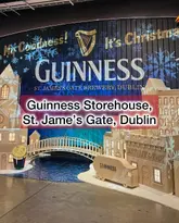 A wide, eye-level shot of a festive, miniature display at the Guinness Storehouse in Dublin, featuring a model of the brewery and a bridge. The image is heavily obstructed by a large, semi-transparent text overlay in the center of the frame.