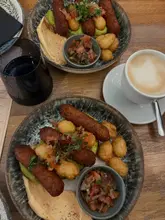 A top-down, casual photograph of two brunch plates featuring veggie sausages, smashed avocado on sourdough, hash browns, and salsa, accompanied by a coffee. The image captures a typical cafe dining experience, focusing on the meal rather than the environment.