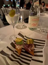 A moody, atmospheric shot taken at a restaurant table, featuring a close-up of a crab-based appetizer on branded paper, accompanied by a martini and a bottle of water. The lighting is warm and intimate, typical of a high-end dining environment.