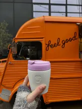 A casual, lifestyle-focused shot featuring a reusable coffee cup held in front of a branded orange food truck. The image captures a moment of post-workout refreshment, effectively using the branded vehicle as a backdrop to establish the venue's identity.