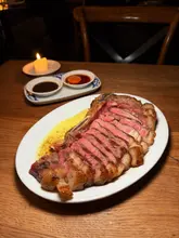 A high-quality, moody food photograph featuring a perfectly cooked, sliced bone-in sirloin steak served with dipping sauces in a dimly lit restaurant setting. The image effectively uses shallow depth of field to draw attention to the texture and doneness of the meat, creating an inviting and sophisticated atmosphere.
