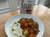A home-cooked meal featuring a tofu and vegetable curry served with rice, presented alongside the retail sauce mix packaging. The shot is a standard, functional food photograph taken in a home environment.