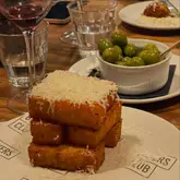A close-up, candid shot of a stack of polenta fries topped with parmesan, served at a restaurant table. The image captures an authentic dining experience with a glass of wine and olives in the background, creating a warm, inviting atmosphere.