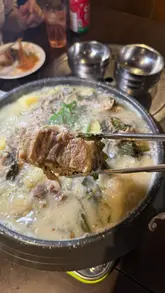 A montage video showcasing a variety of Korean dishes at Jin Go Gae restaurant, including pork bone soup, Japchae, beef stir-fry, marinated crab, and fried chicken. The video features close-up shots of the food and a group toast, set to music with text overlays identifying the dishes.