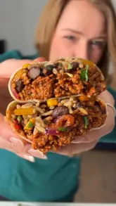 A short, instructional video demonstrating how to assemble a vegan tempeh wrap. The creator shows the layering of ingredients including rice, corn, beans, and seasoned tempeh, finishing with a bite of the final product.