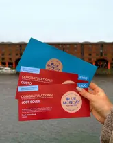 A promotional photograph featuring a hand holding two 'Blue Monday' gift vouchers in front of the Royal Albert Dock in Liverpool. The image is designed to announce a giveaway event, with the vouchers clearly visible and the location providing immediate context for the promotion.