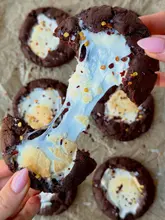 A high-quality, close-up food photography shot capturing the 'pull' of a melted marshmallow inside a chocolate cookie. The lighting is bright and even, highlighting the texture of the melted marshmallow and the dark chocolate cookie, with a shallow depth of field that keeps the focus on the central action while providing context with other cookies in the background.