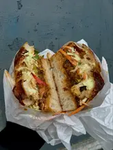 A close-up, top-down shot of a fried sweet potato and tofu sandwich in a paper wrapper, held in someone's hand. The sandwich is cut in half, revealing the filling.