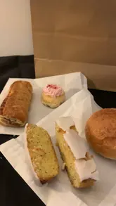 A creator picks up a 'Too Good To Go' bag from Bread and Brew in Wendover, showing the contents including a bread roll, lemon drizzle cake, Eton mess cake, and a vegan sausage roll.