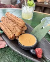 A close-up, appetizing shot of churros served with a dipping sauce, a single blueberry, and a raspberry on a dark plate, held by a person. The background features a blurred drink, suggesting a relaxed dining or social setting.