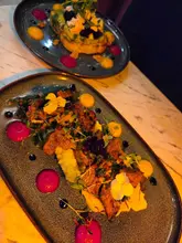 A high-angle, close-up shot of two dishes served at a restaurant. The foreground features a plate with steak, mashed potatoes, chimichurri, and edible flowers, while the background shows a plate of bruschetta with goat cheese, beetroot, and avocado.