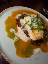 A close-up, top-down shot of a Cornish hake dish served with Sicilian caponata, sea herbs, and a clam butter sauce, presented on a ceramic plate.