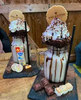 A close-up, eye-level shot of two elaborate 'freakshakes' served on slate boards, featuring whipped cream, brownies, and various chocolate bars, set against a rustic wooden wall background.