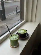 A simple, candid shot of two matcha drinks—one hot and one iced—placed on a windowsill, capturing a quiet moment in a cafe setting.