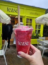 A bright, eye-level shot of a vibrant pink smoothie held in front of a distinctive yellow shop front at Bicester Village. The branding on the cup is clear, and the natural lighting highlights the texture of the drink.